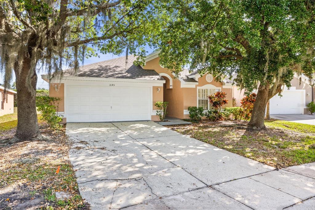 A tan single-story house with a two-car garage and a concrete driveway under large trees — 3145 STONEHURST CIRCLE, KISSIMMEE, FL