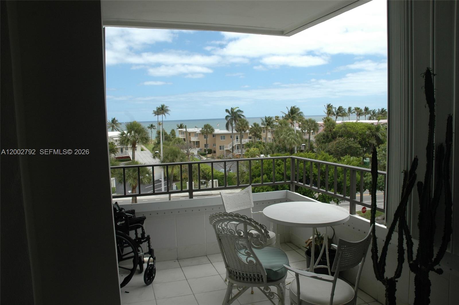 A balcony with patio furniture and a wheelchair overlooks a coastal view with palm trees — 1401 S Ocean Blvd # 408, Pompano Beach FL 33062, POMPANO BEACH, FL