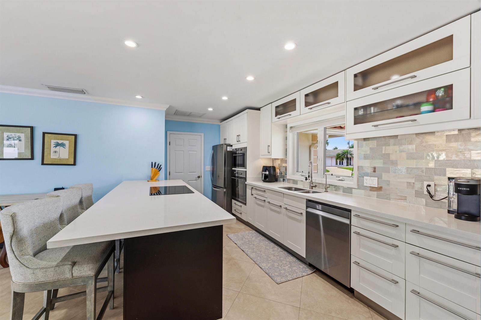 A modern kitchen features white cabinetry, a dark island with a cooktop, and light-blue walls — 1205 Dolphin Road, Singer Island FL 33404, SINGER ISLAND, FL