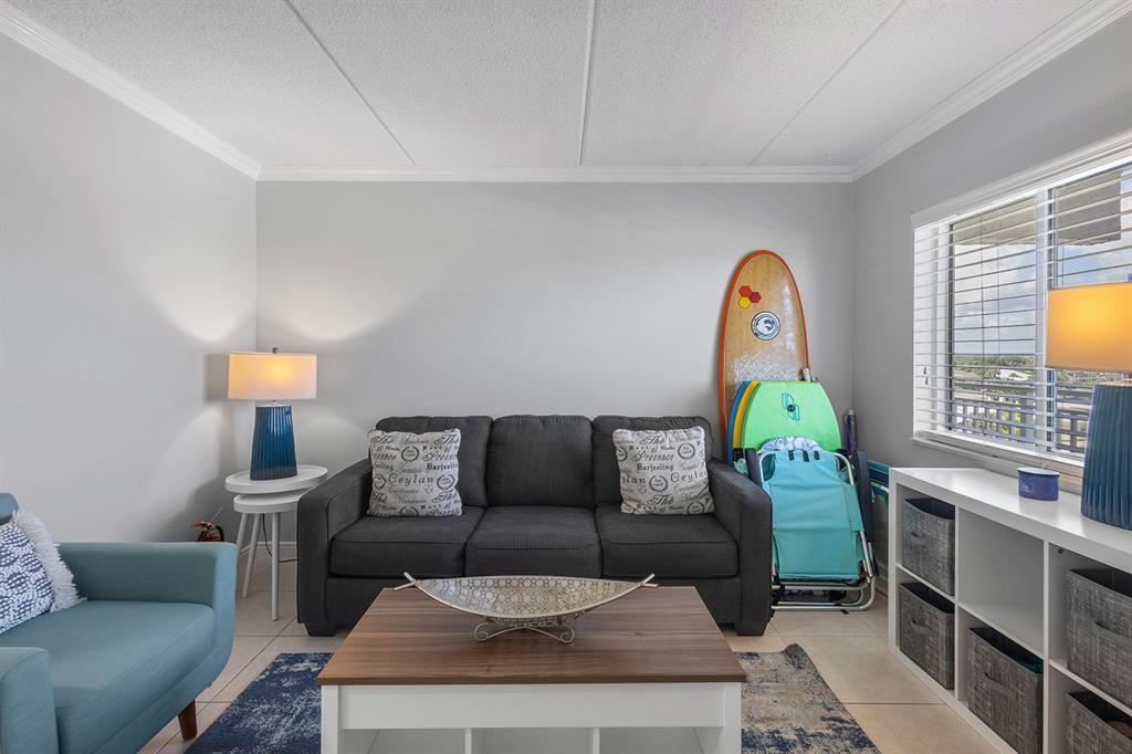 A living room features a dark gray sofa, a coffee table, and a surfboard leaning against — 2401 S ATLANTIC AVENUE UNIT A603, NEW SMYRNA BEACH, FL