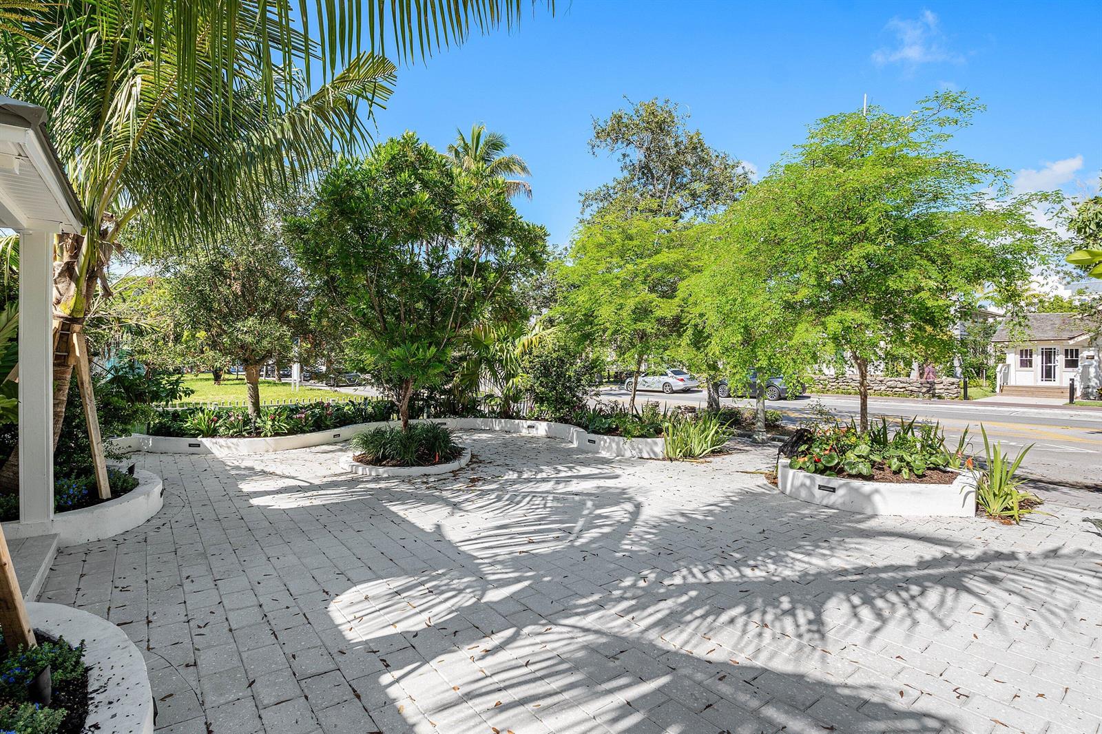 A paved patio area features curved garden beds with trees and shrubs under a blue sky — 23 S Swinton Avenue, Delray Beach FL 33444, DELRAY BEACH, FL