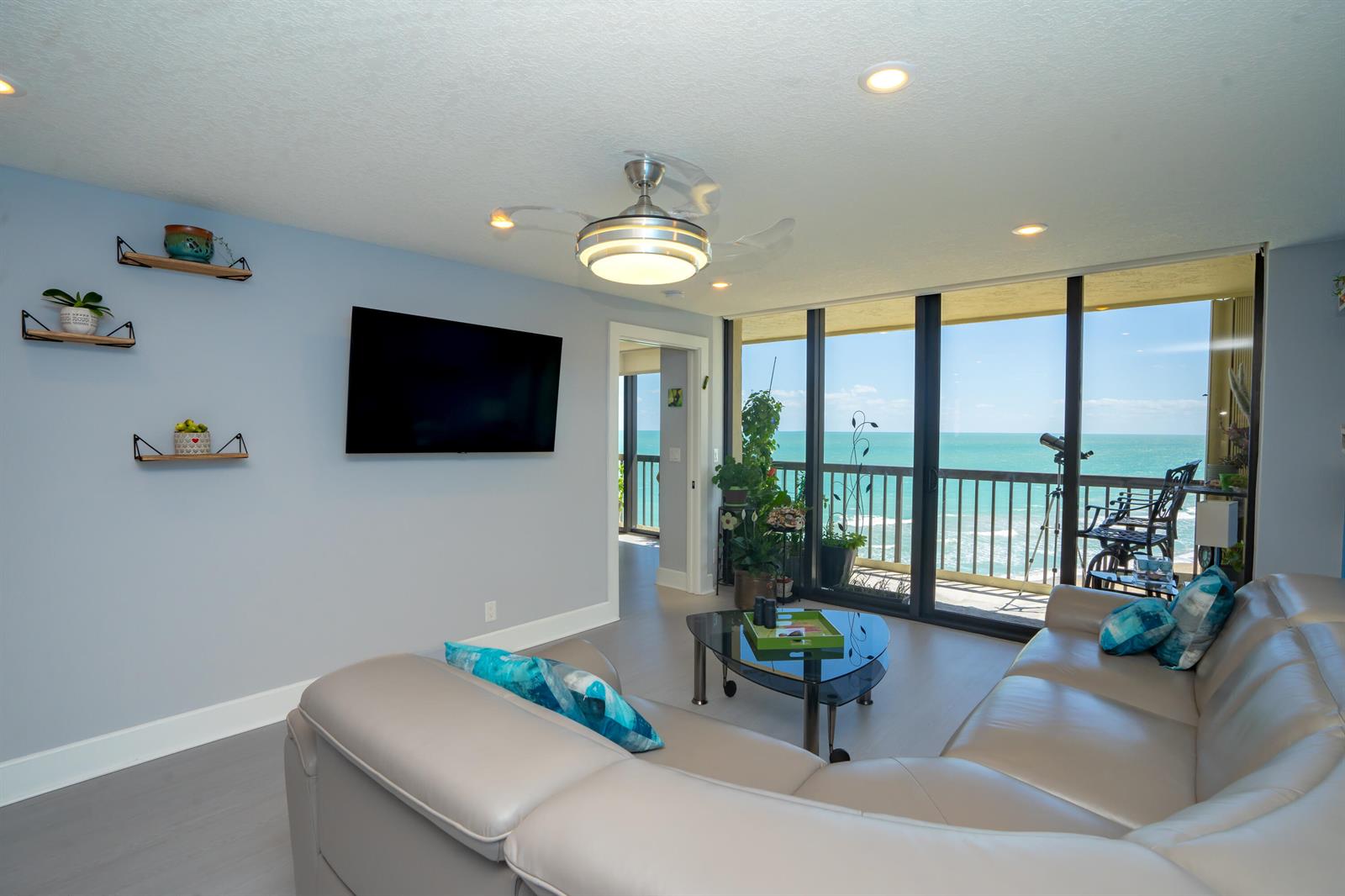 A living room features a light gray sectional, wall-mounted television, and floor-to-ceiling windows overlooking the ocean — 9500 S Ocean Drive # 1208, Jensen Beach FL 34957, JENSEN BEACH, FL