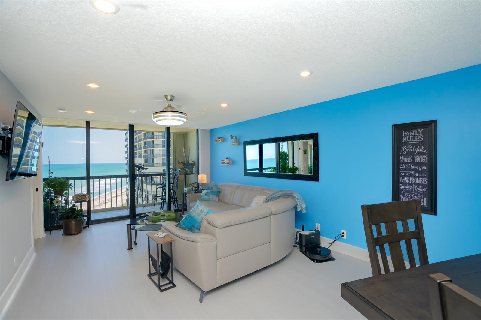 A living room with a light-colored sofa, blue accent wall, and a balcony overlooking the ocean — 9500 S Ocean Drive # 1208, Jensen Beach FL 34957, JENSEN BEACH, FL