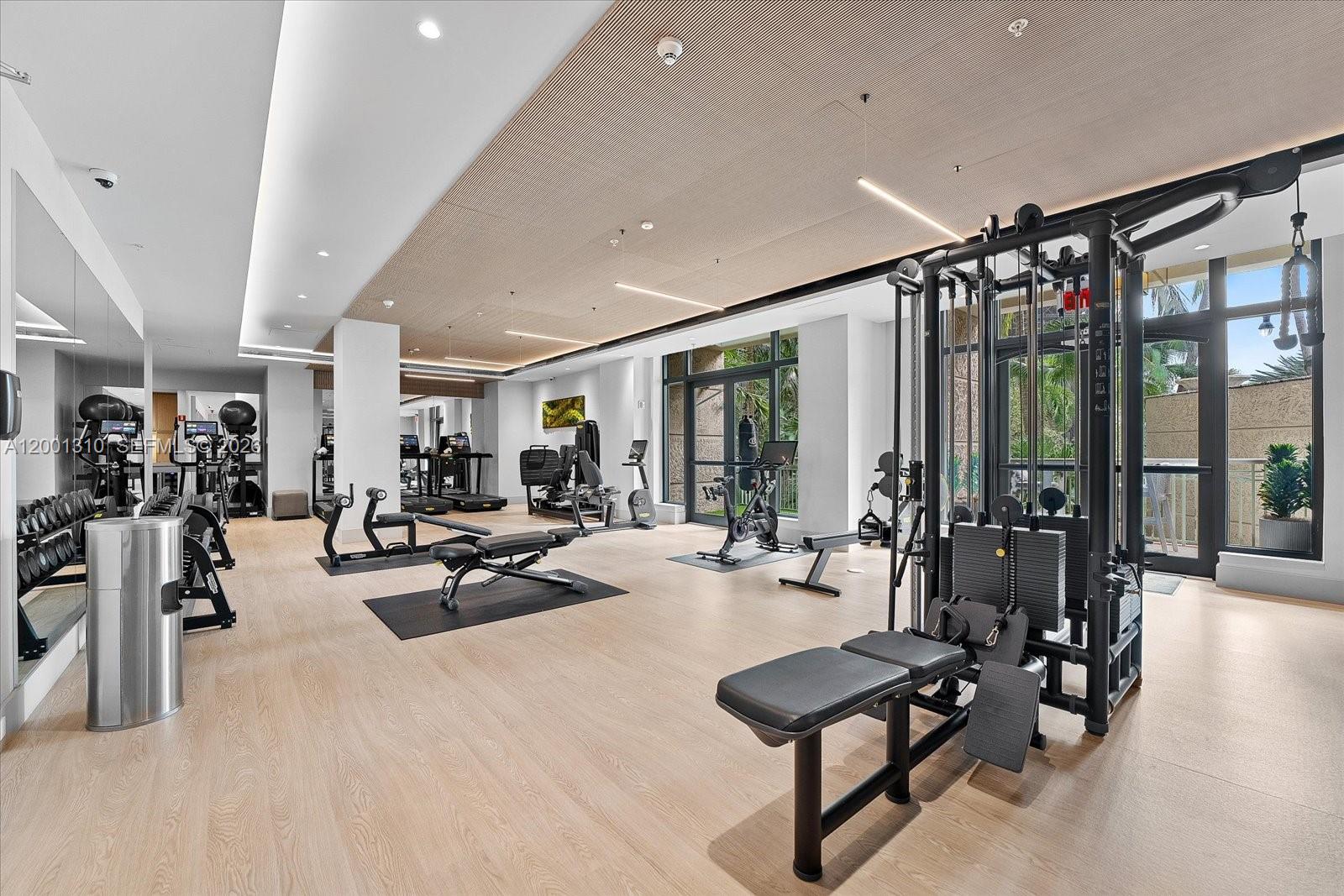 A modern fitness center features light wood flooring, various exercise machines, and floor-to-ceiling windows — 3400 SW 27th Ave # 201, Miami FL 33133, MIAMI, FL