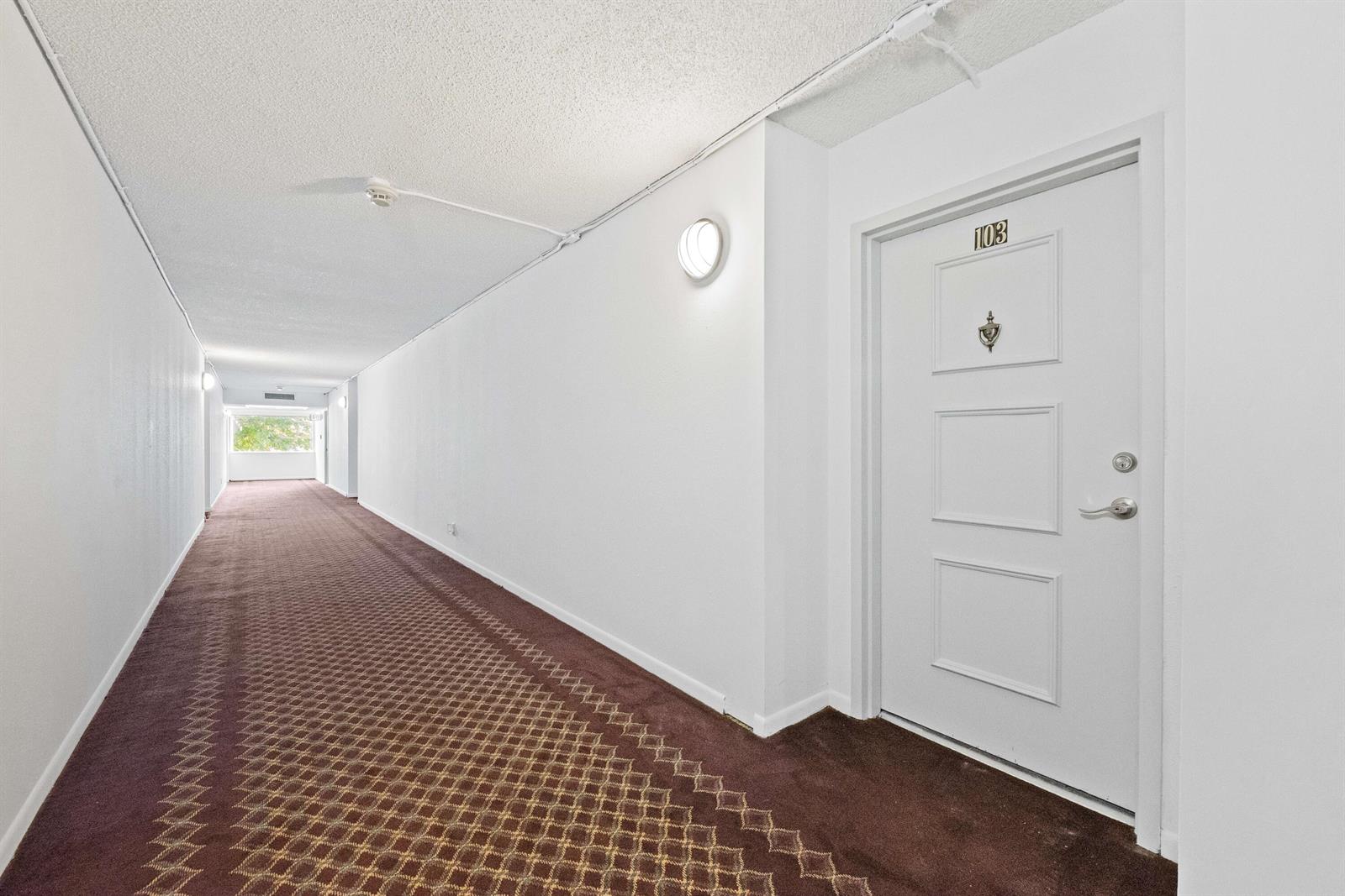 A long, carpeted apartment hallway features a white door labeled 103 and wall-mounted lighting — 140 Lakeview Drive # 103, Weston FL 33326, WESTON, FL