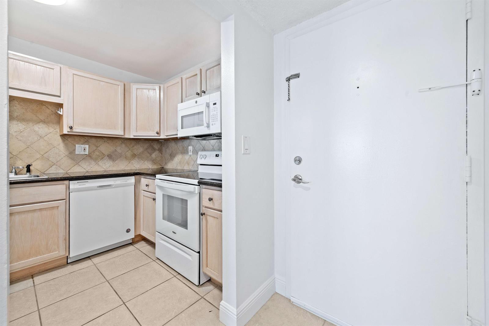A kitchen features light wood cabinets, white appliances, a tiled backsplash, and a white door — 140 Lakeview Drive # 103, Weston FL 33326, WESTON, FL