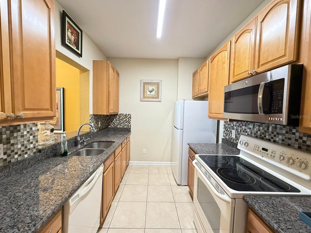 A galley kitchen features light wood cabinets, granite countertops, a tiled backsplash, and white appliances — 6466 CAVA ALTA DRIVE UNIT 108, ORLANDO, FL