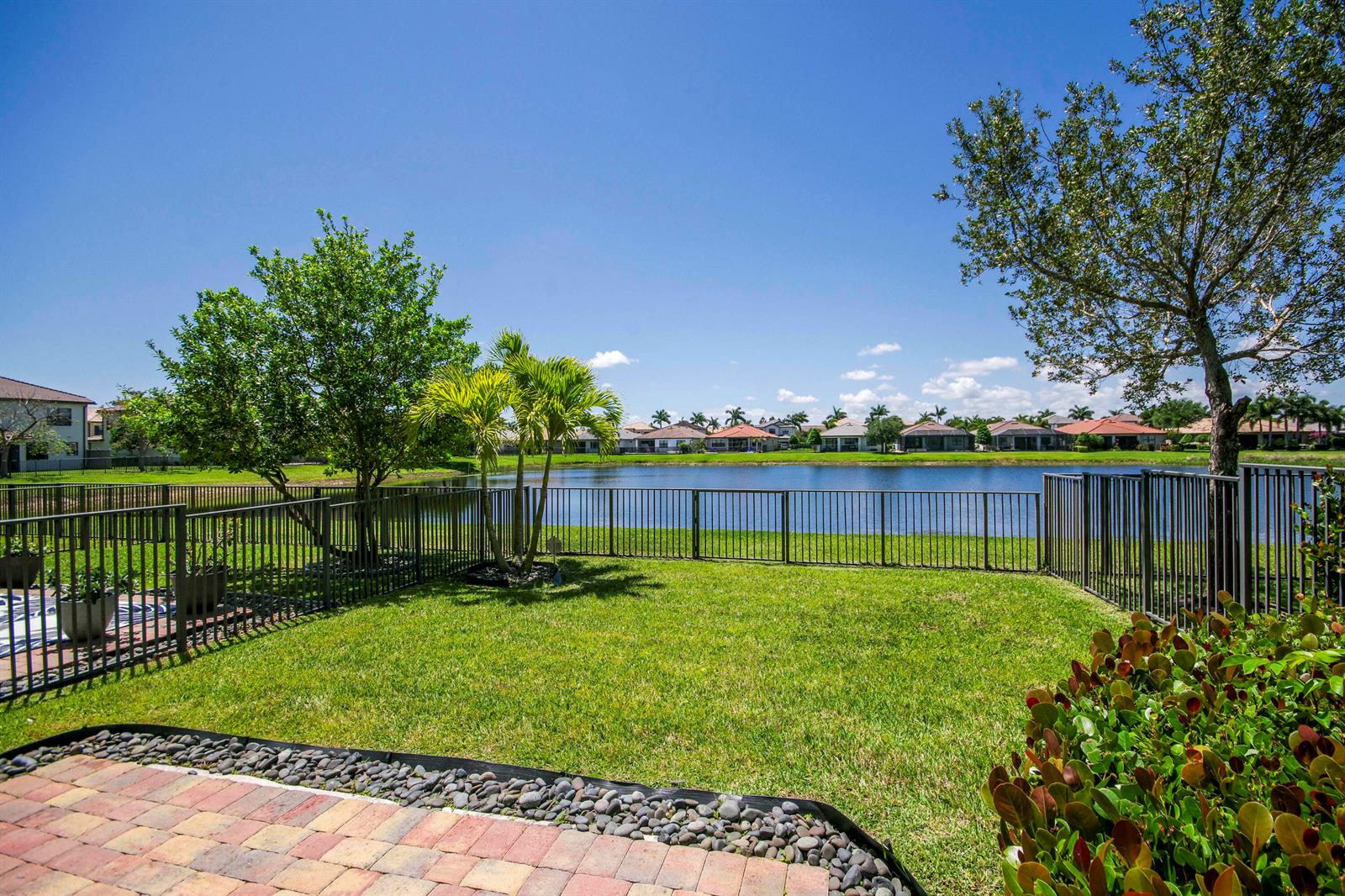 A grassy backyard features a metal fence, palm trees, and a view of a lake — 8301 Rearing Lane, Lake Worth FL 33467, LAKE WORTH, FL