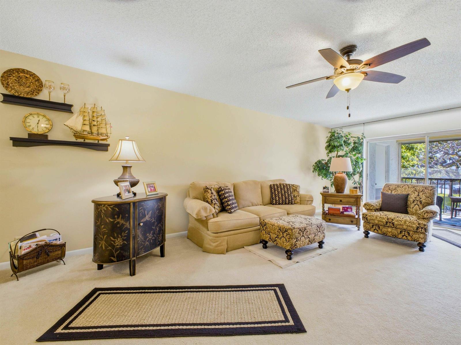 A living room features a beige sofa, patterned armchair, ottoman, and wall-mounted shelves with decor — 6625 S Oriole Boulevard # 2040, Delray Beach FL 33446, DELRAY BEACH, FL