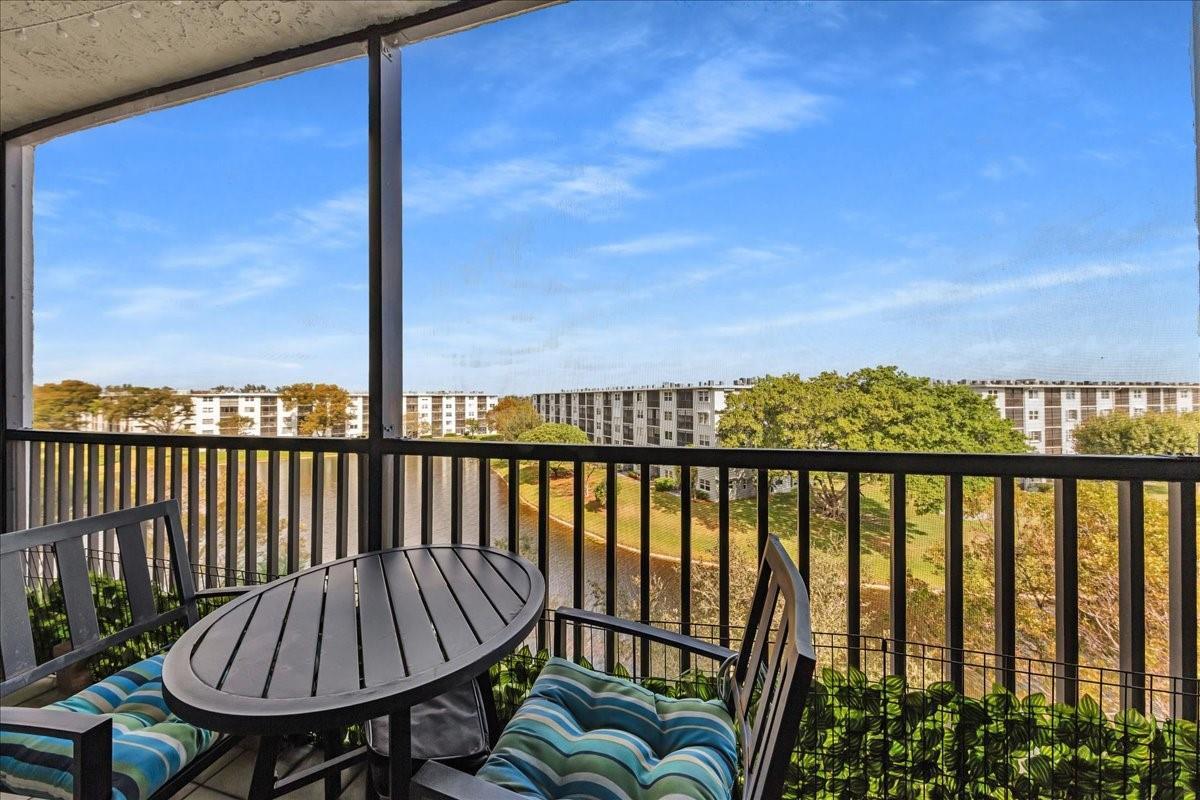 A screened-in balcony features a small table and chairs overlooking a pond and apartment buildings — 2217 Cypress Island Drive # 507, Pompano Beach FL 33069, POMPANO BEACH, FL