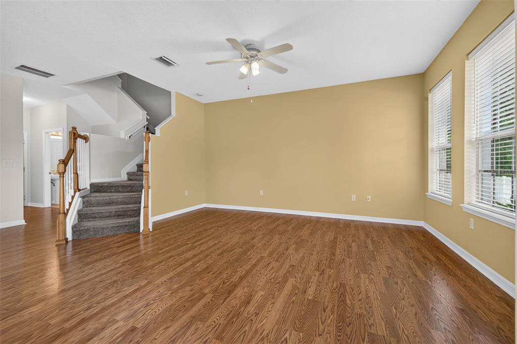 An empty living room features tan walls, wood-look flooring, a ceiling fan, and a staircase — 27747 PLEASURE RIDE LOOP, WESLEY CHAPEL, FL