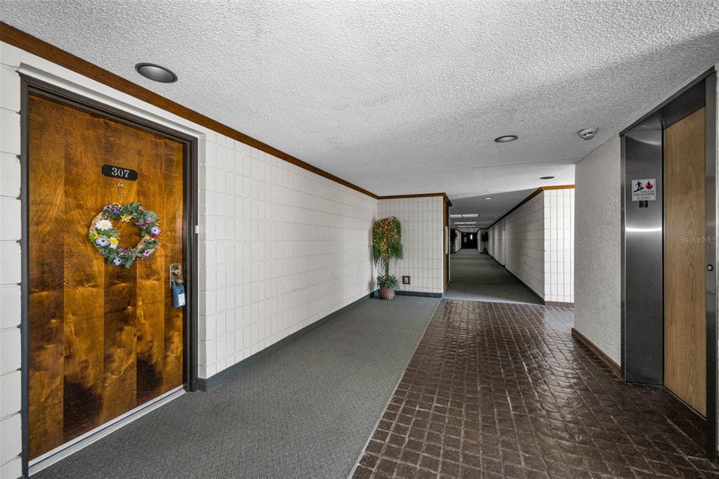 A hallway features a wooden door with a wreath, tiled walls, and an elevator entrance — 13612 S VILLAGE DRIVE UNIT 5307, TAMPA, FL