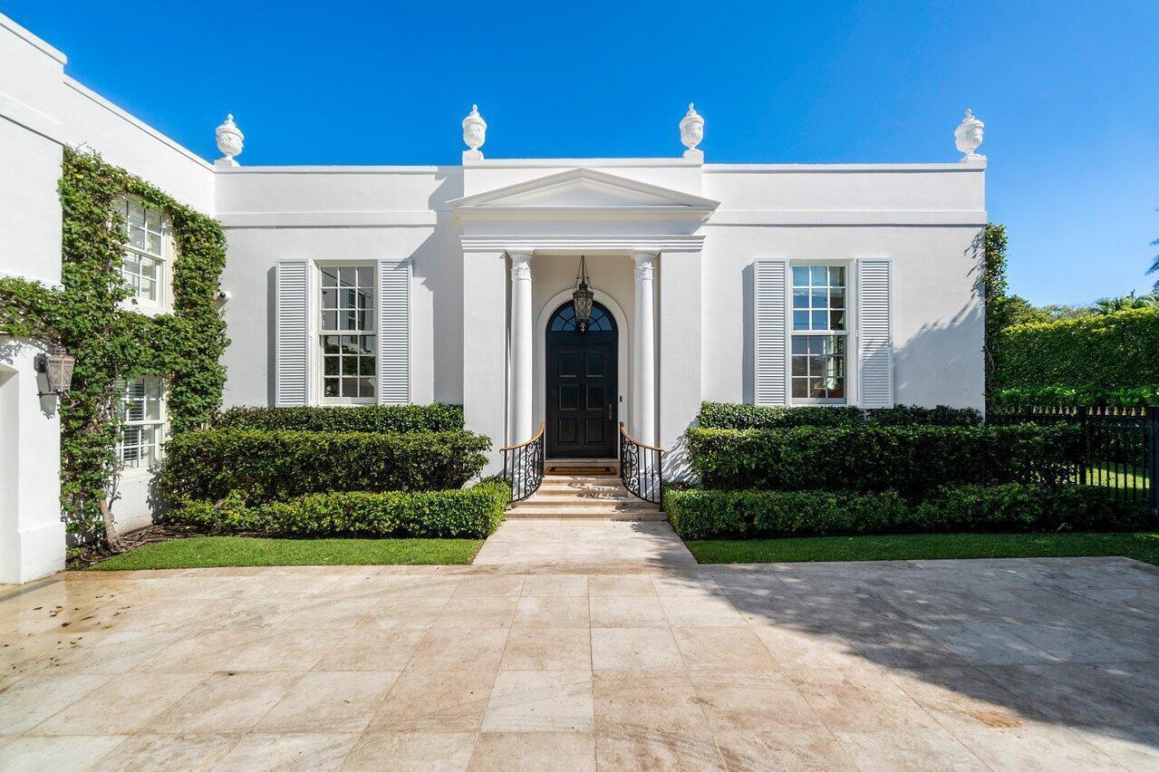 A white neoclassical house features a dark front door, columns, and symmetrical windows with shutters — 100 Regents Park Road, Palm Beach FL 33480, PALM BEACH, FL