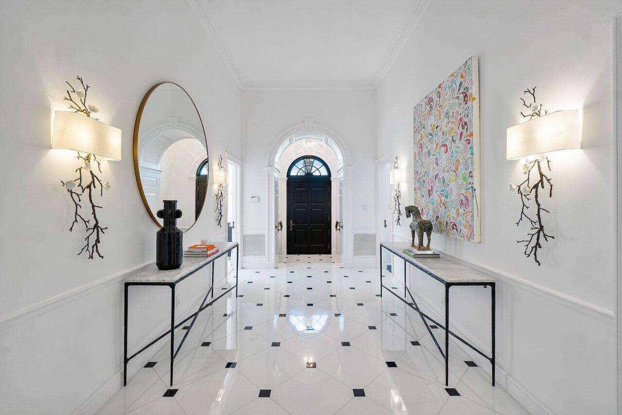 A symmetrical foyer features white marble floors with black accents, two console tables, and wall sconces — 100 Regents Park Road, Palm Beach FL 33480, PALM BEACH, FL