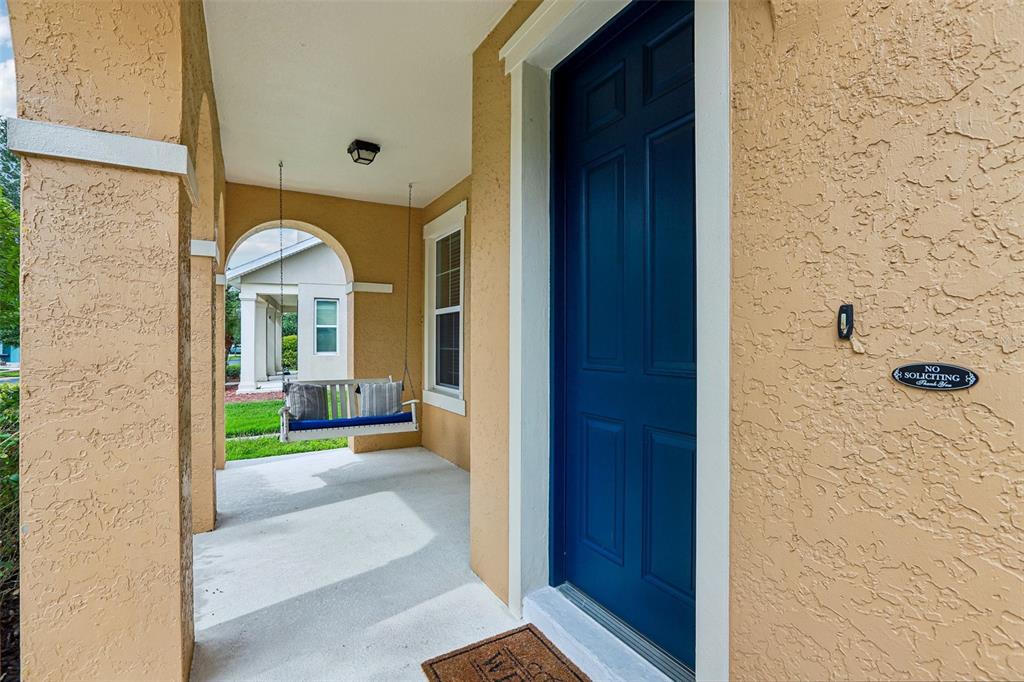 A covered front porch features a blue door, a hanging bench, and stucco walls — 5233 SUNCATCHER DRIVE, WESLEY CHAPEL, FL