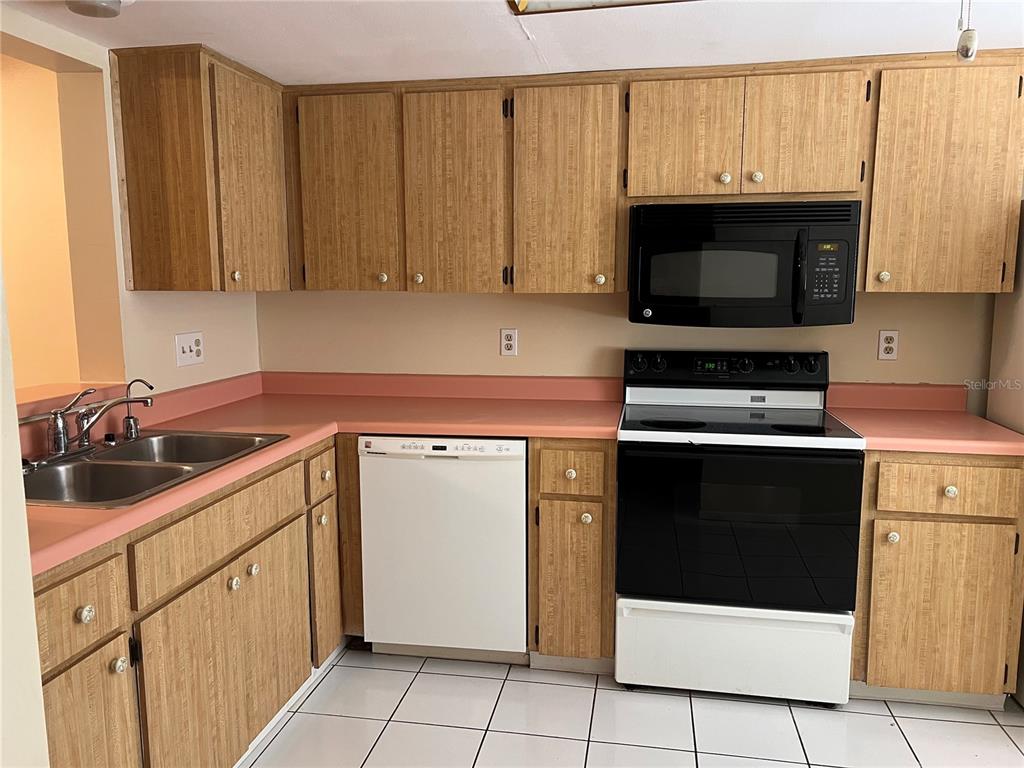 A kitchen features light wood cabinets, pink laminate countertops, white appliances, and white floor tiles — 2225 LARK CIRCLE W UNIT E, PALM HARBOR, FL
