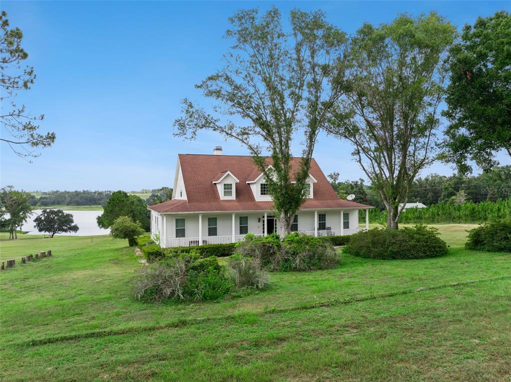 A white two-story farmhouse with a red roof sits on a grassy hill near water — 25895 TILLY HAWK LANE, BROOKSVILLE, FL