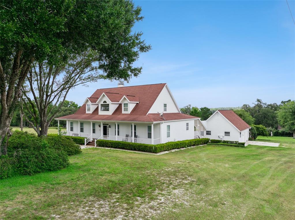 A white two-story farmhouse with a red roof and a wraparound porch sits on grass — 25895 TILLY HAWK LANE, BROOKSVILLE, FL
