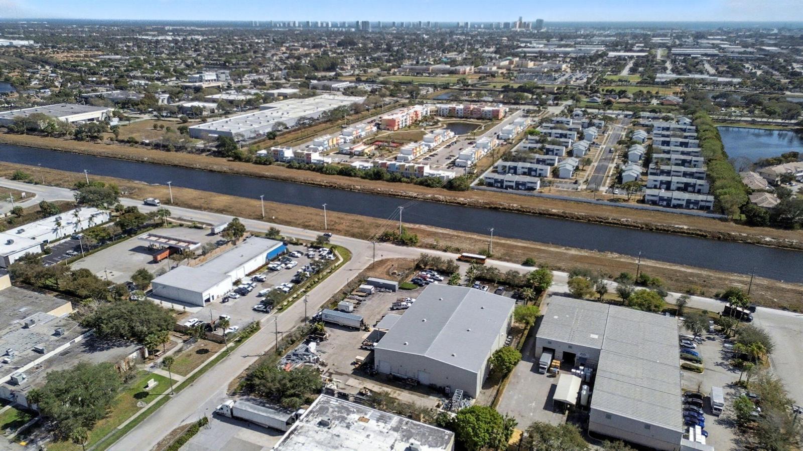 An aerial view shows industrial warehouses, a canal, and residential townhomes in a suburban landscape — 6861 Garden Road, Riviera Beach FL 33404, RIVIERA BEACH, FL