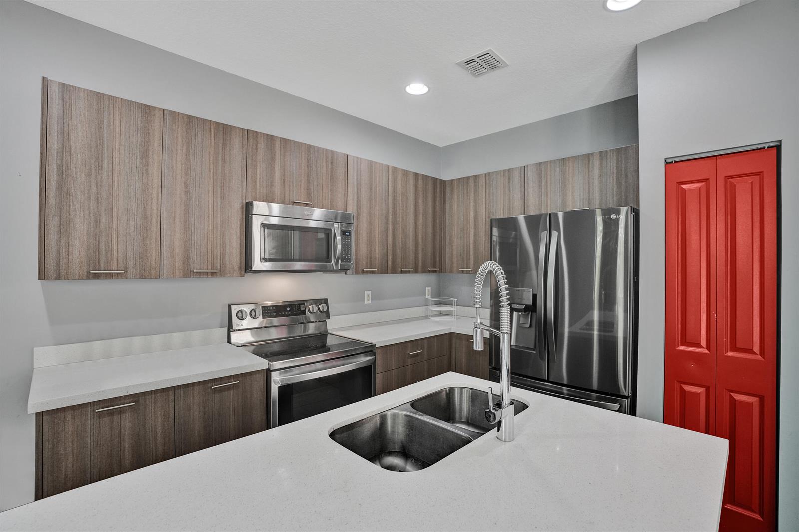 A modern kitchen features wood-grain cabinets, stainless steel appliances, white countertops, and a red door — 5902 NW 46th Lane, Tamarac FL 33319, TAMARAC, FL