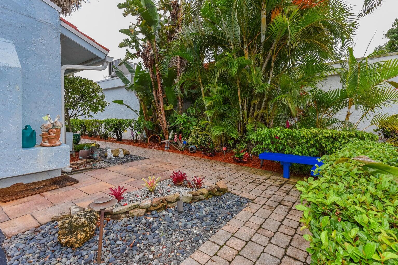 A paved patio area features a blue bench, lush tropical landscaping, and decorative stone accents — 5518 Ainsley Court, Boynton Beach FL 33437, BOYNTON BEACH, FL