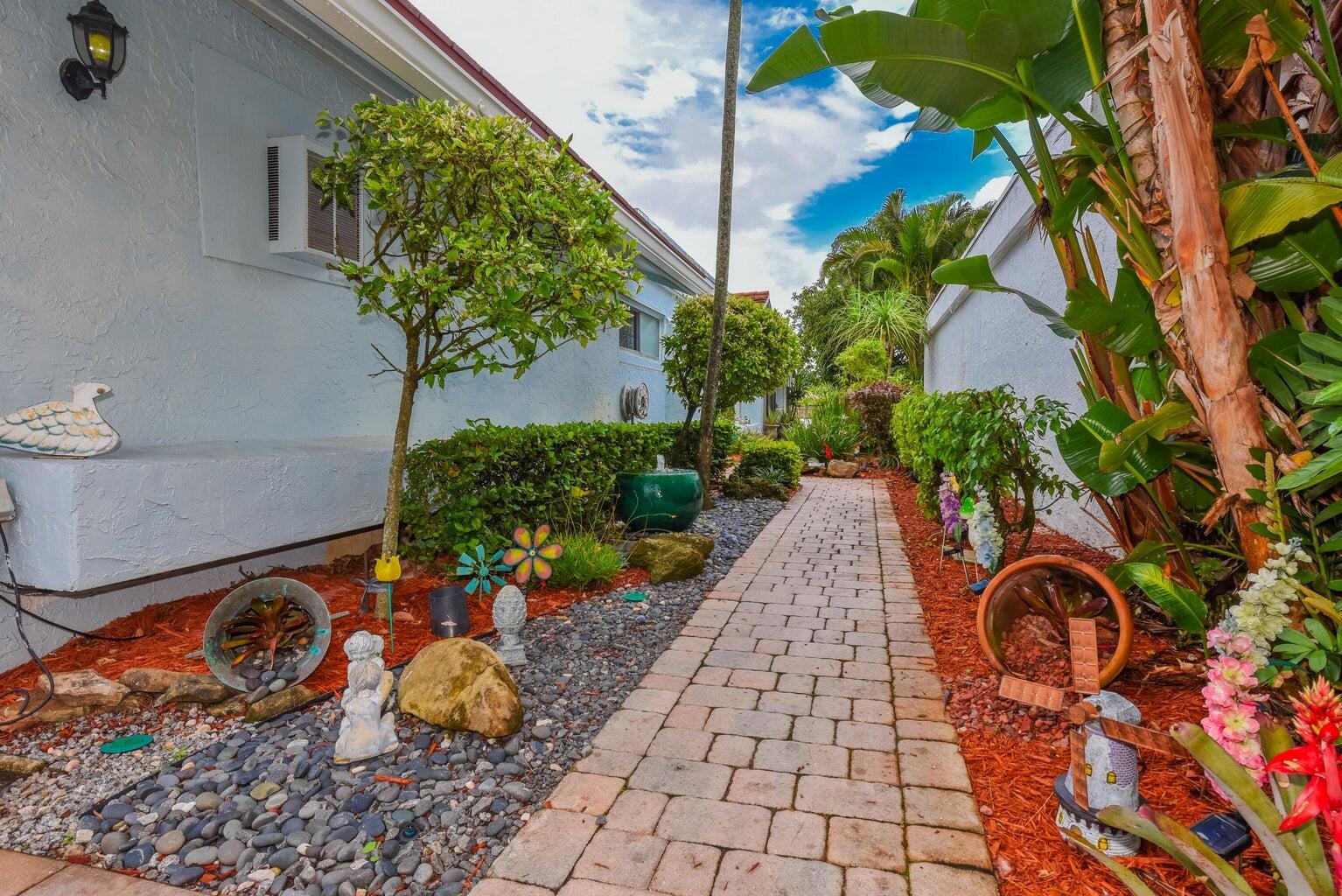 A paved walkway leads between two light blue exterior walls surrounded by landscaping and garden decor — 5518 Ainsley Court, Boynton Beach FL 33437, BOYNTON BEACH, FL