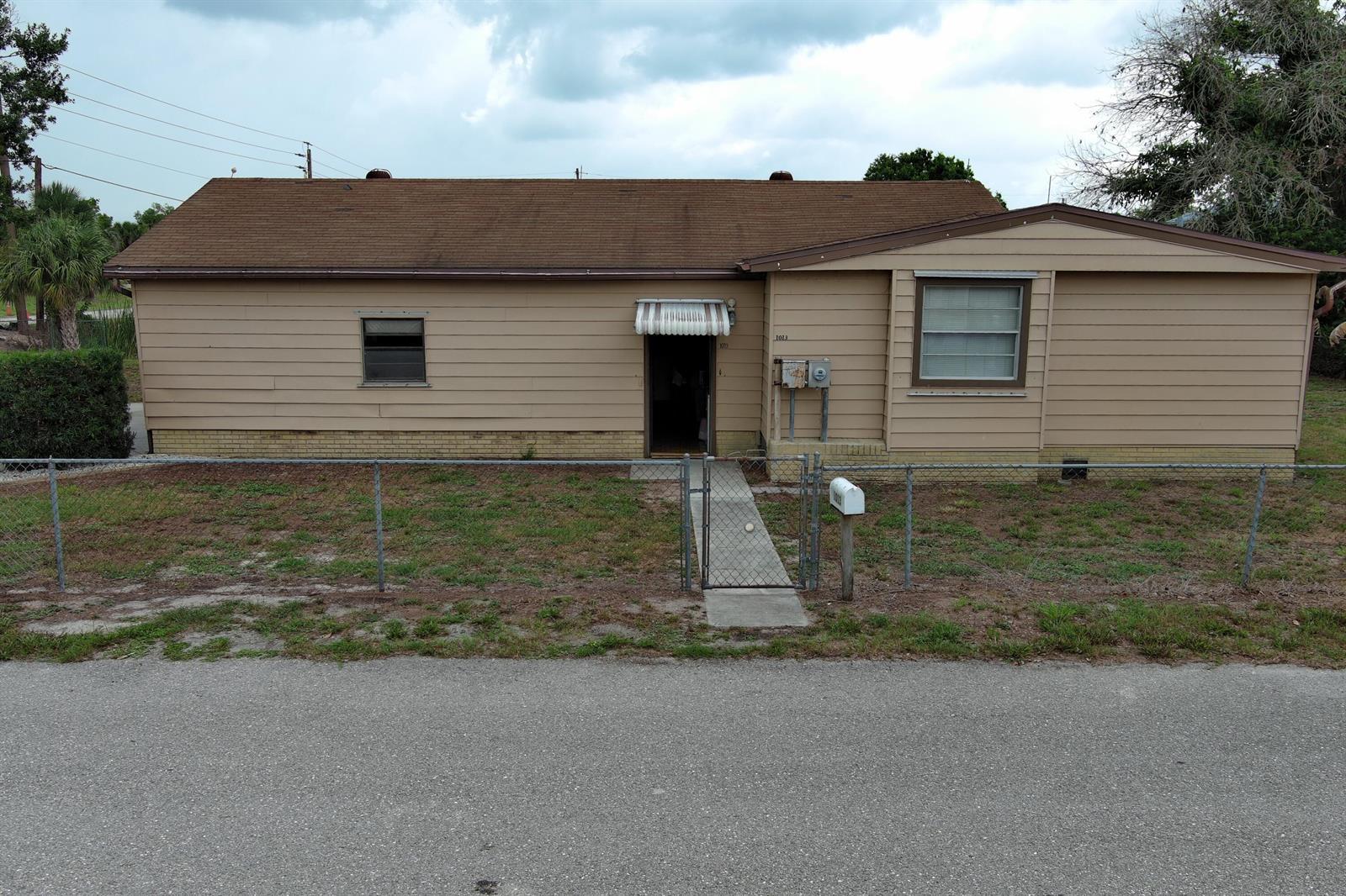 A single-story tan house with a brown roof sits behind a chain-link fence — 1013 9th Street, Okeechobee FL 34974, OKEECHOBEE, FL