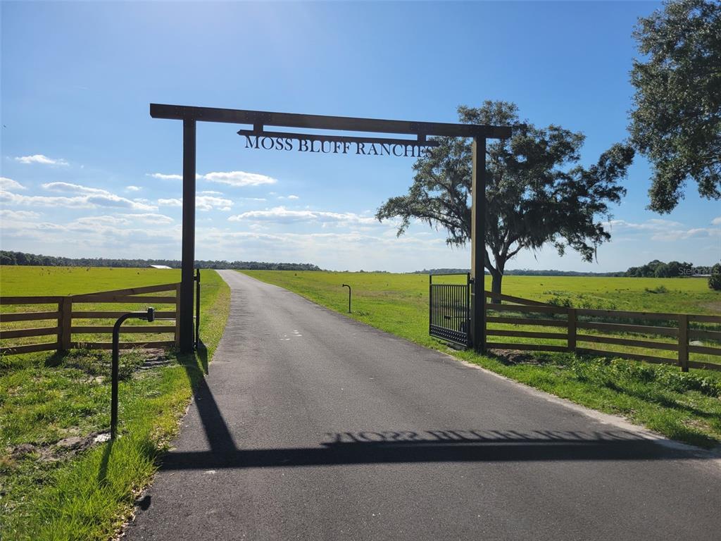 An asphalt driveway leads through a wooden entrance gate labeled Moss Bluff Ranches — TBD SE 170 AVE ROAD UNIT Tract A, OCKLAWAHA, FL