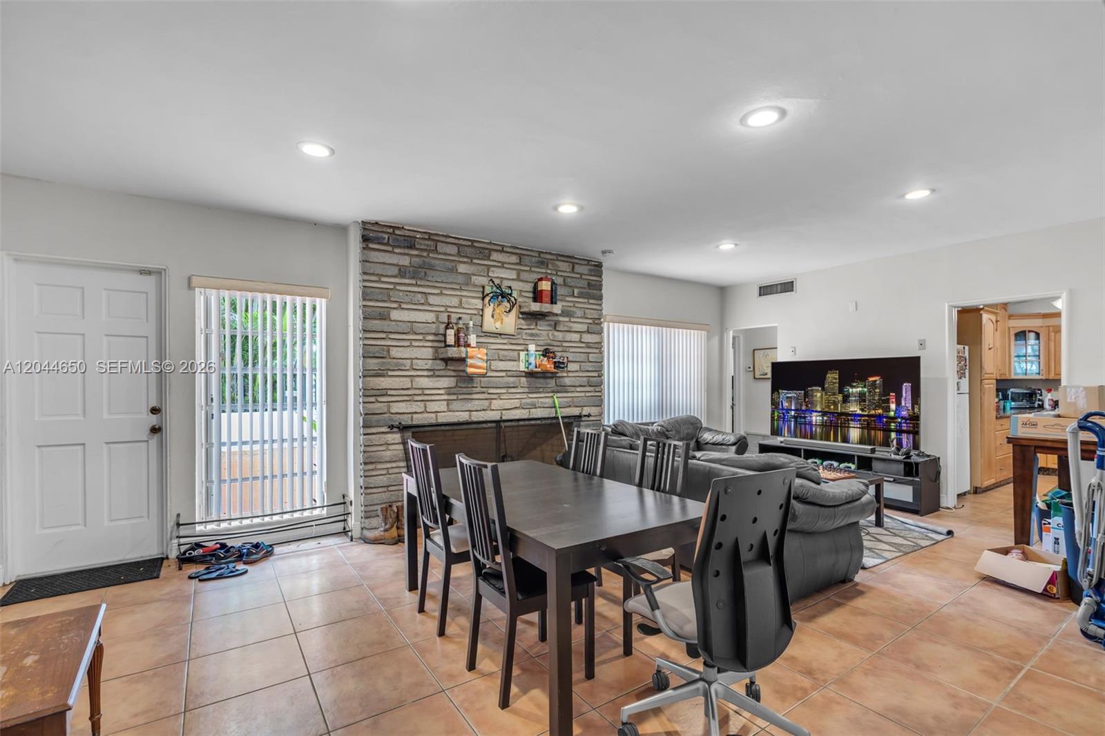 A dining area with a brick fireplace wall and tile flooring opens into a living room — 1439 Miller Rd, Coral Gables FL 33146, CORAL GABLES, FL
