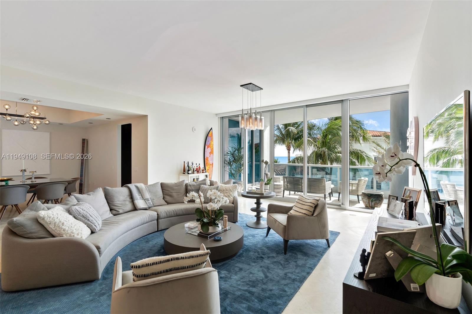 A living room with a curved sofa, blue rug, and floor-to-ceiling windows overlooking a balcony — 5959 Collins Ave # 606, Miami Beach FL 33140, MIAMI BEACH, FL