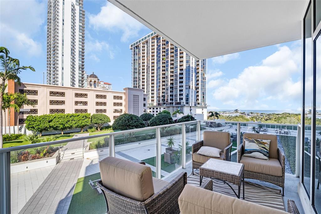 A balcony with wicker furniture overlooks a courtyard, surrounding high-rise buildings, and the distant ocean — 175 1ST STREET S UNIT 702, ST PETERSBURG, FL