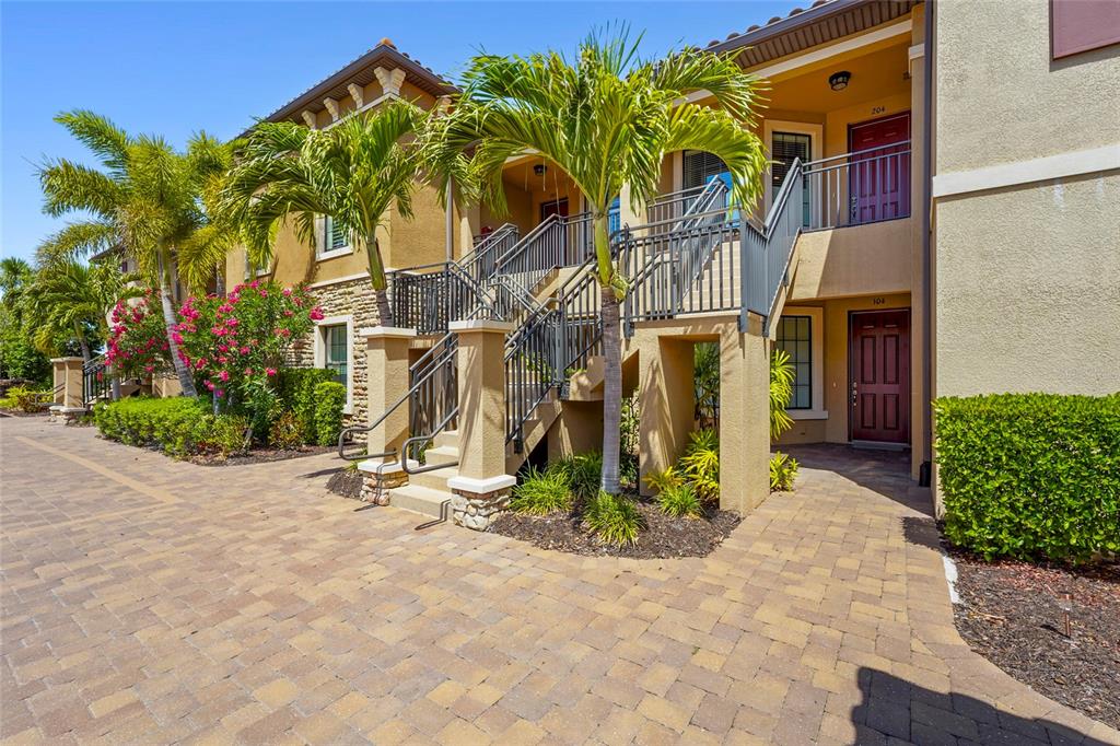 A two-story apartment building exterior features a stone-accented staircase, palm trees, and paved walkways — 5420 CICERNONE STREET UNIT 203, SARASOTA, FL