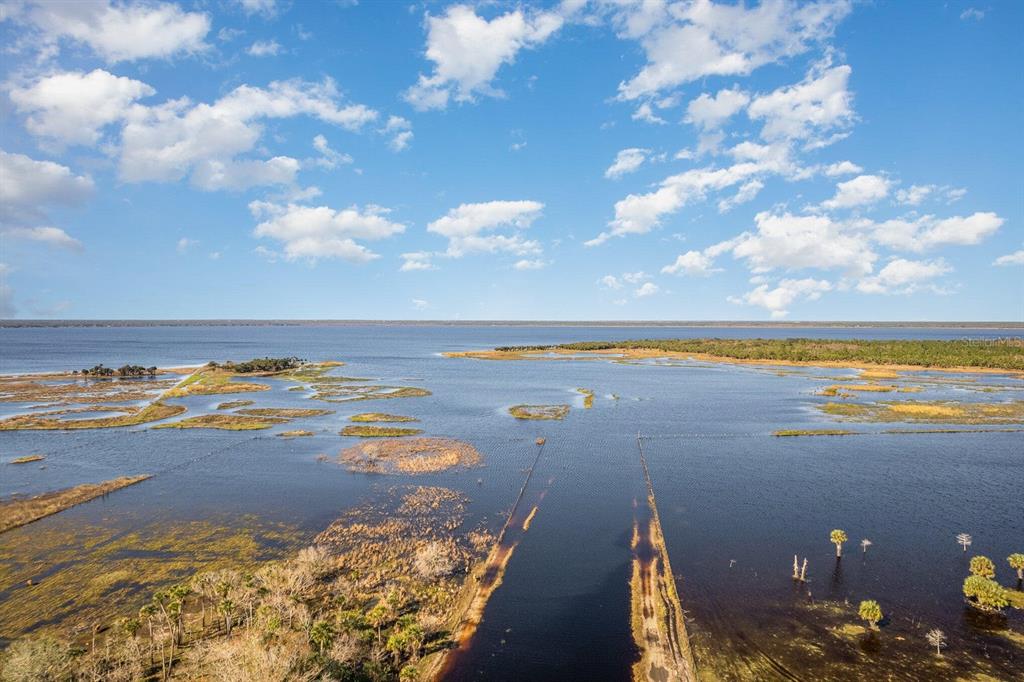 An aerial view shows a large lake with marshy islands and a flooded dirt path — LAKE HARNEY WOODS BOULEVARD, OSTEEN, FL