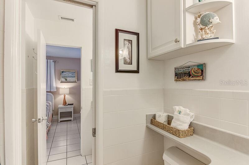 A bathroom with white tiled walls, a wicker basket, and a doorway leading to a bedroom — 6020 MIDNIGHT PASS ROAD UNIT 1, SARASOTA, FL