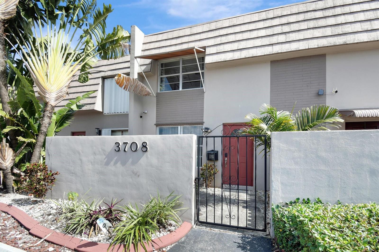 A two-story townhouse exterior features a gated courtyard, a red front door, and tropical landscaping — 3708 SW 59th Terrace # 68, Davie FL 33314, DAVIE, FL