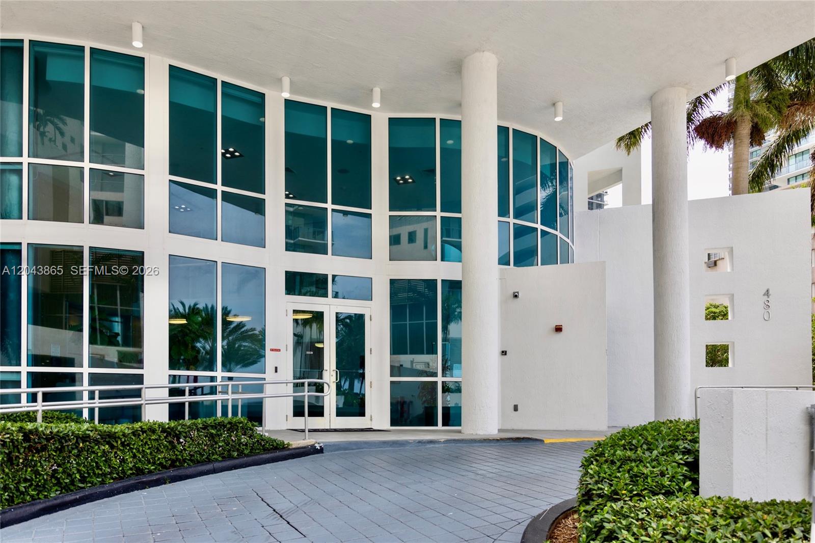 The exterior entrance of a modern building features glass windows, white columns, and a driveway — 480 NE 30th St # 1507, Miami FL 33137, MIAMI, FL