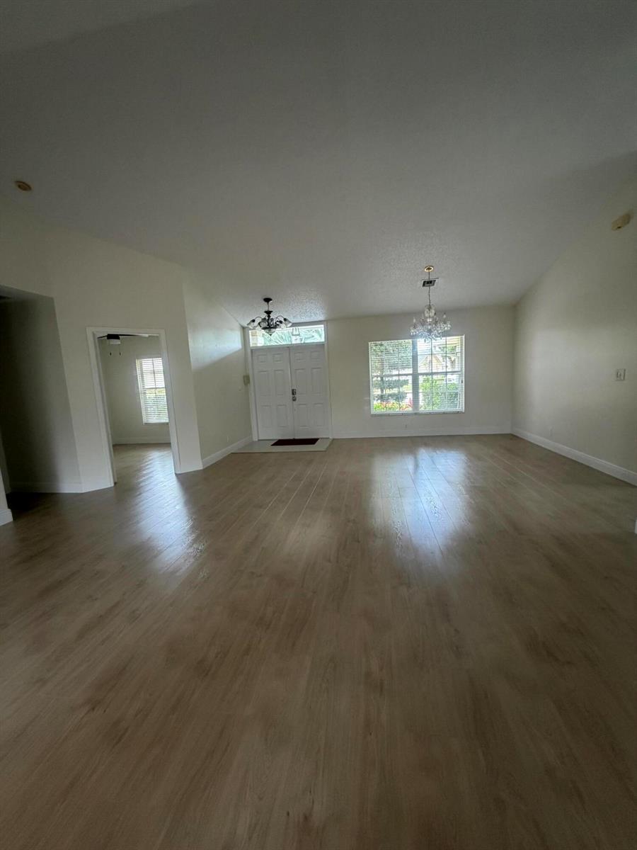 A spacious, empty living room features light wood-look flooring, white walls, and two chandeliers — 1401 Hideaway Bend, Wellington FL 33414, WELLINGTON, FL