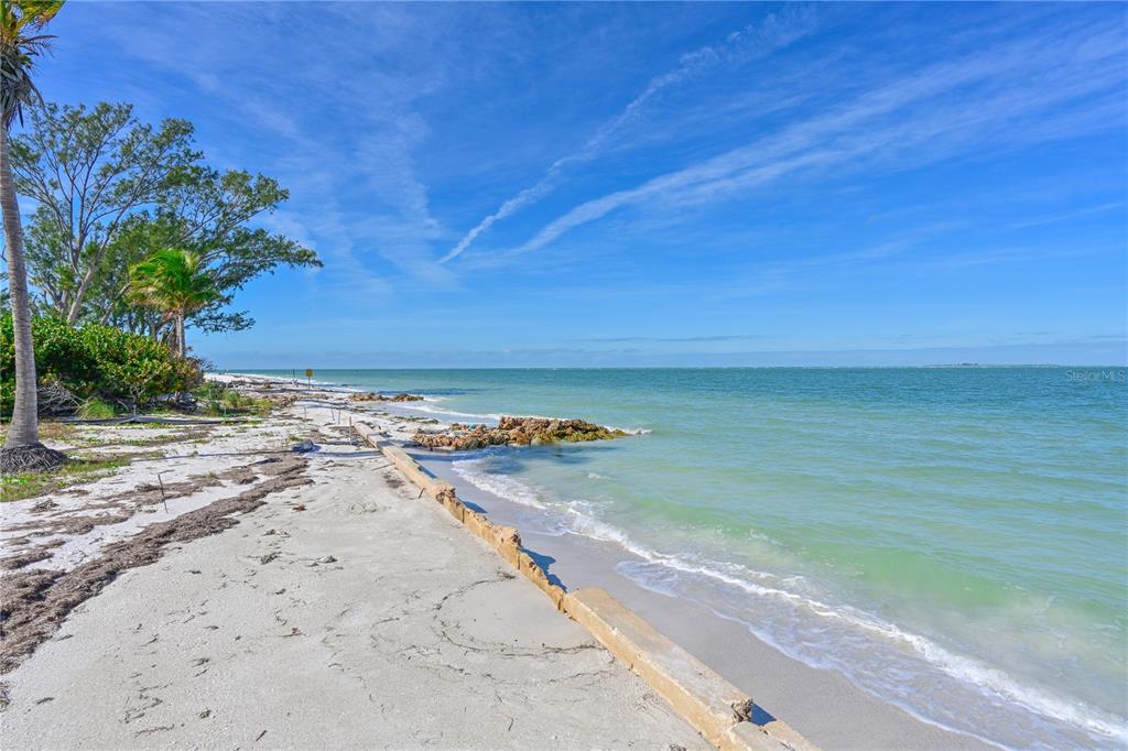 A sandy beach shoreline features a low concrete retaining wall and scattered rocks by water — 839 N SHORE DRIVE, ANNA MARIA, FL