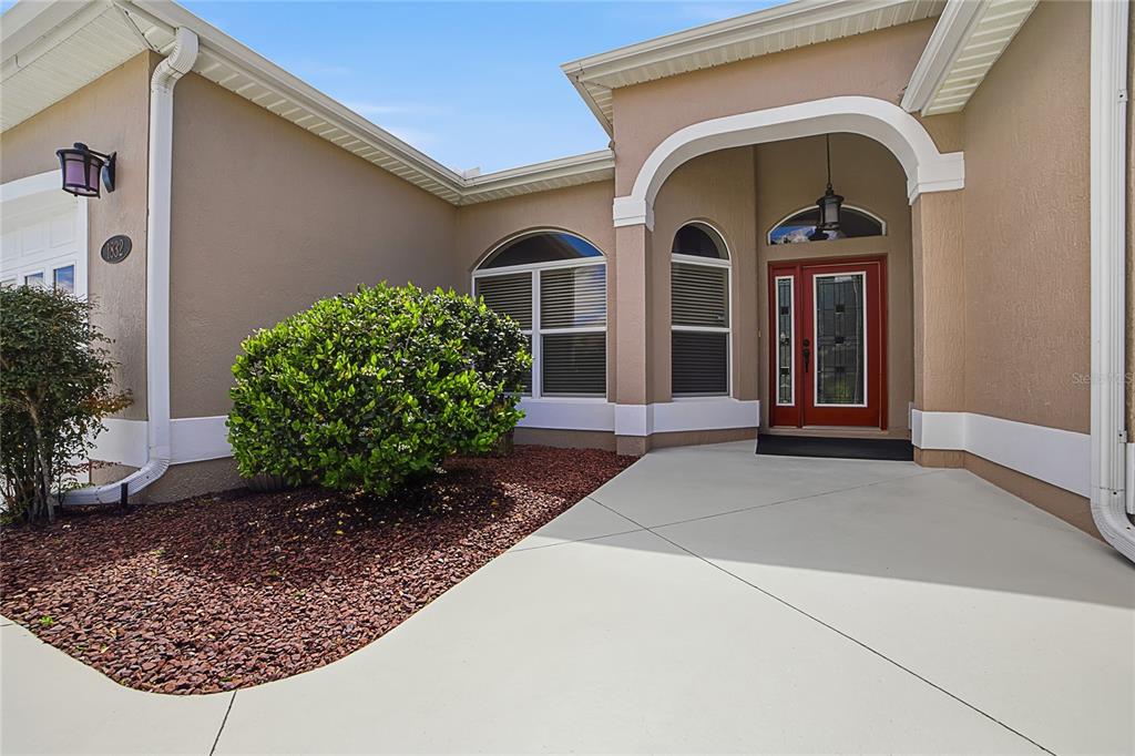 A tan house exterior features a red front door, arched windows, and a concrete entryway — 1832 HARTFORD PATH, THE VILLAGES, FL