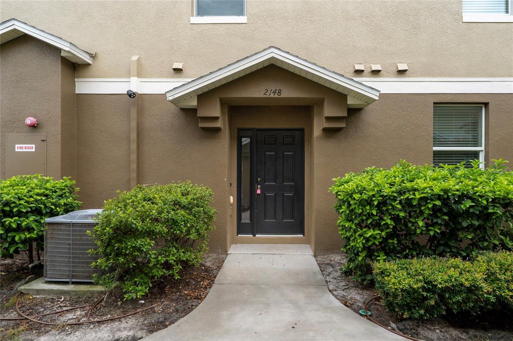 A concrete walkway leads to the dark front door of a tan stucco building — 2148 BETSY ROSS LANE UNIT 2148, ST CLOUD, FL