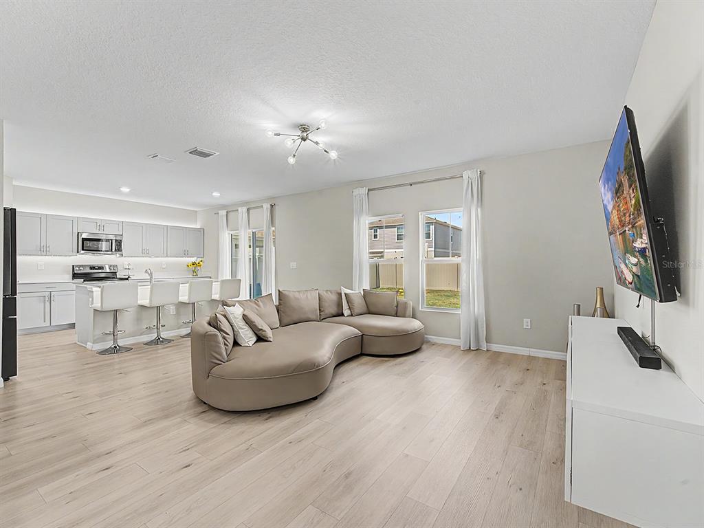 An open-concept living room and kitchen featuring light wood flooring and a curved sectional sofa — 7587 STARFLOWER WAY, WESLEY CHAPEL, FL