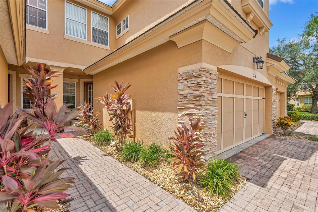 A tan two-story house exterior features a stone-accented garage, a brick walkway, and tropical landscaping — 8228 VILLA GRANDE COURT, SARASOTA, FL