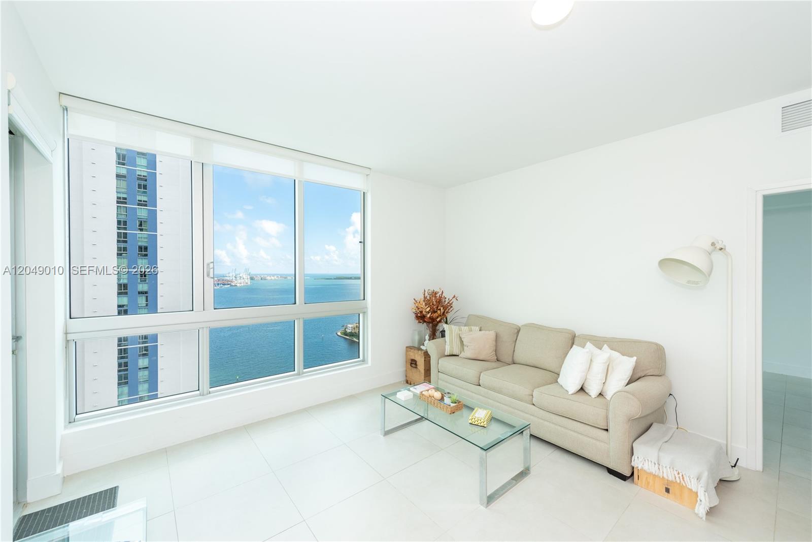 A living room features a beige sofa, glass coffee table, and large windows overlooking water — 300 S Biscayne Blvd # 2814, Miami FL 33131, MIAMI, FL