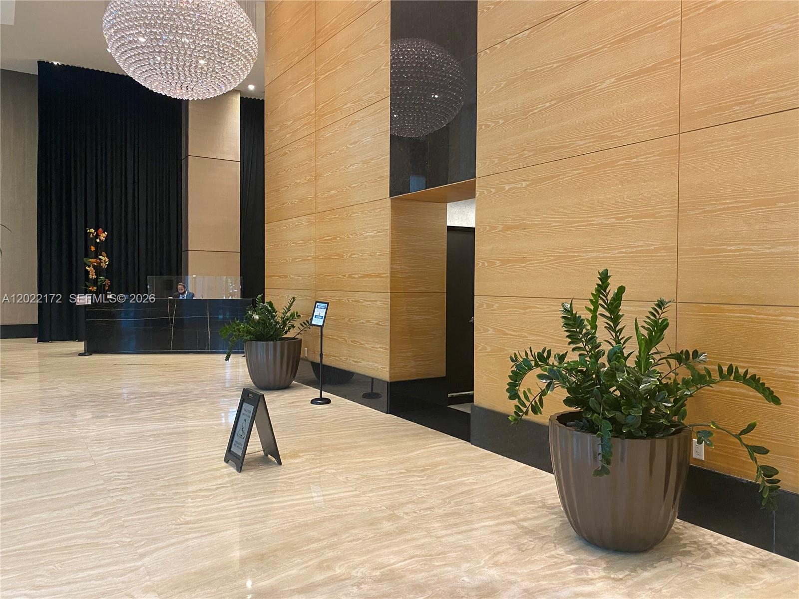 A modern lobby features a reception desk, wood-paneled walls, potted plants, and a crystal chandelier — 950 Brickell Bay Dr # 2206, Miami FL 33131, MIAMI, FL