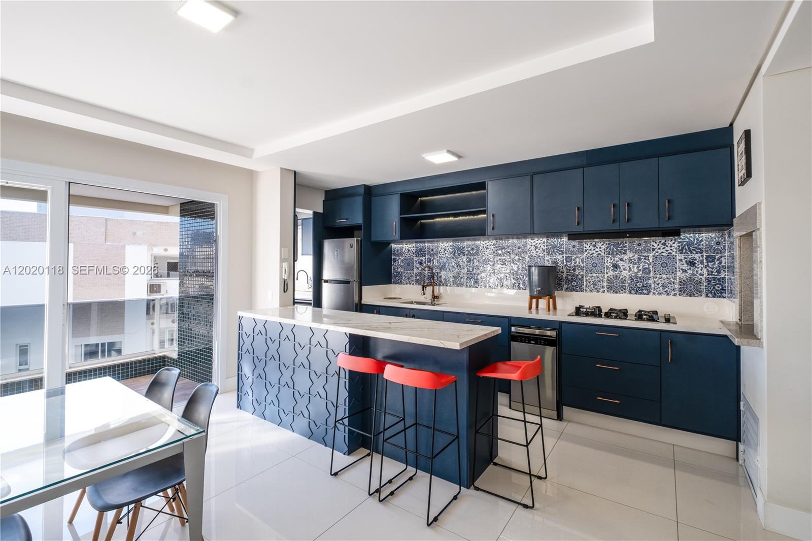 A modern kitchen features dark blue cabinetry, a patterned island, and red bar stools — 1540 Rua das Piraunas # 403, SC 88053418, SC