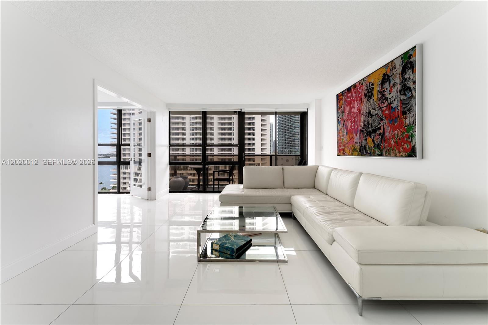 A bright living room features white tile floors, a white sectional sofa, and colorful wall art — 600 NE 36th St # 1111, Miami FL 33137, MIAMI, FL