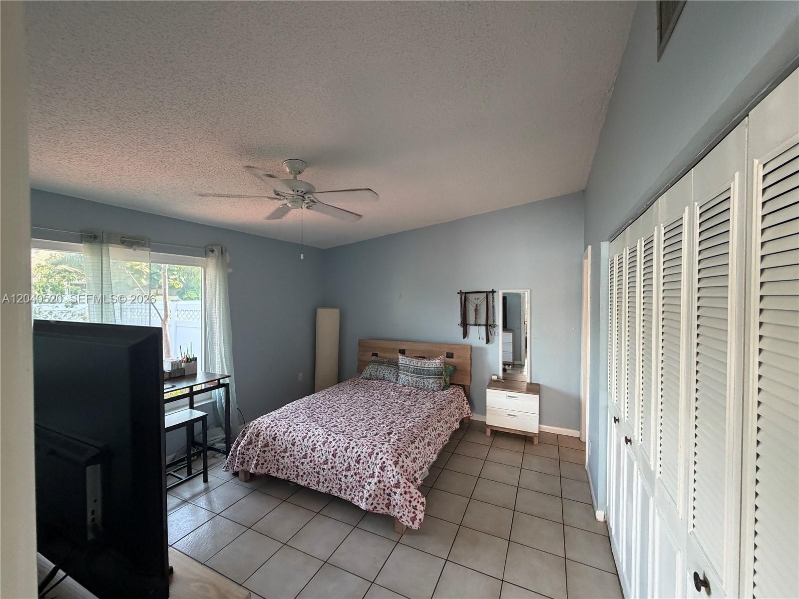 A bedroom features light blue walls, tile flooring, a bed, and white louvered closet doors — 360 B Lakewood Cir E, Margate FL 33063, MARGATE, FL
