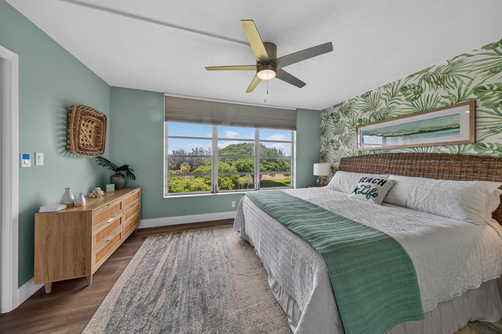 A bedroom features a palm-leaf accent wall, a wicker headboard, and a view of trees — 2650 S Course Drive # 603, Pompano Beach FL 33069, POMPANO BEACH, FL