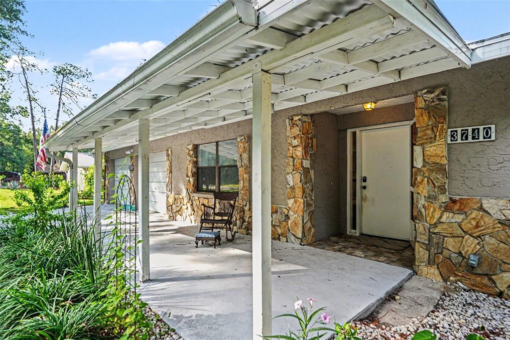 A covered concrete porch features stone accents, a white front door, and a rocking chair — 3740 S EASTPARK WAY, HOMOSASSA, FL