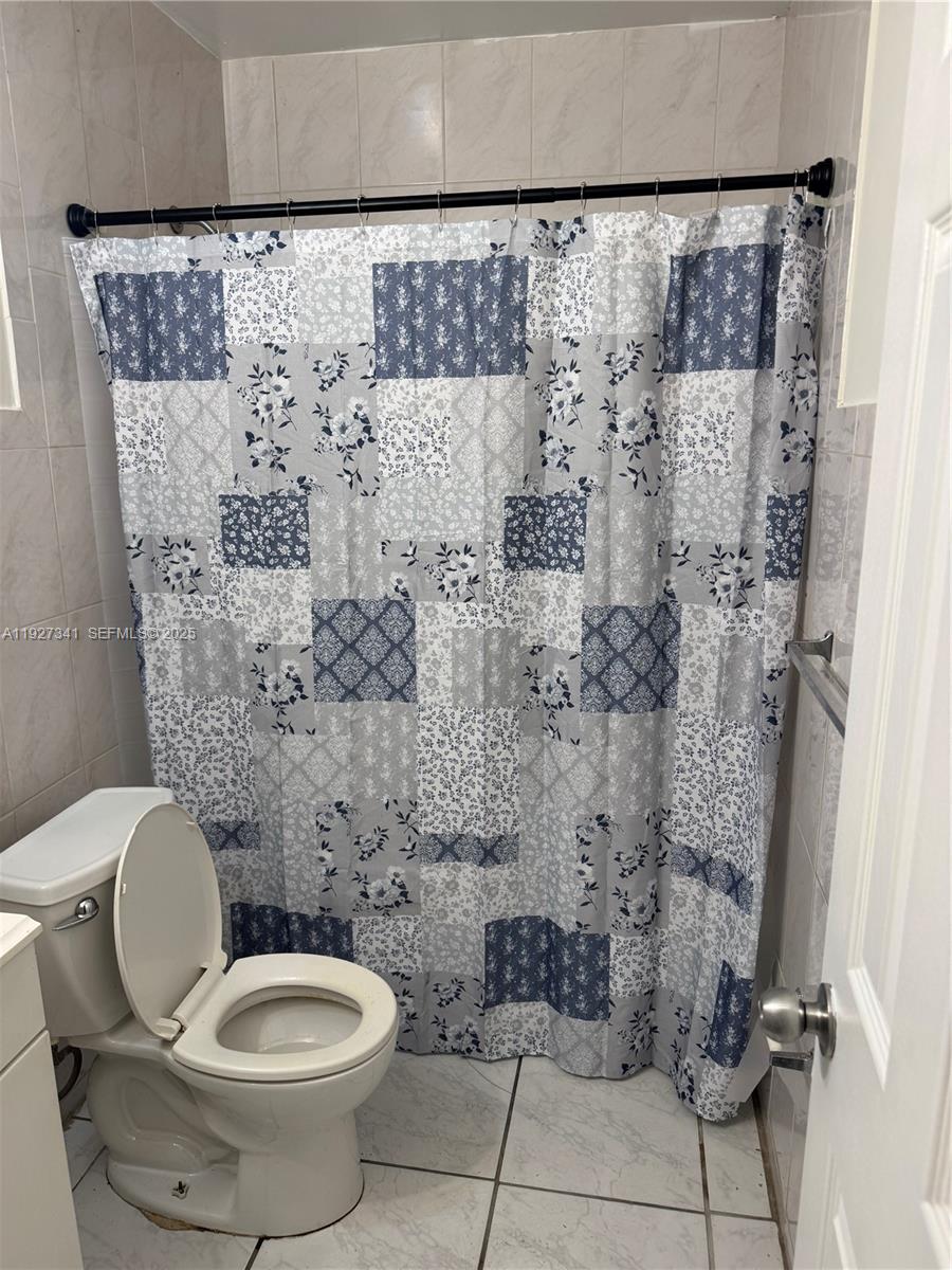A bathroom features a patchwork floral shower curtain, a white toilet, and light-colored tiled walls — 4717 SW 33rd Ave # 202, Dania Beach FL 33312, DANIA BEACH, FL